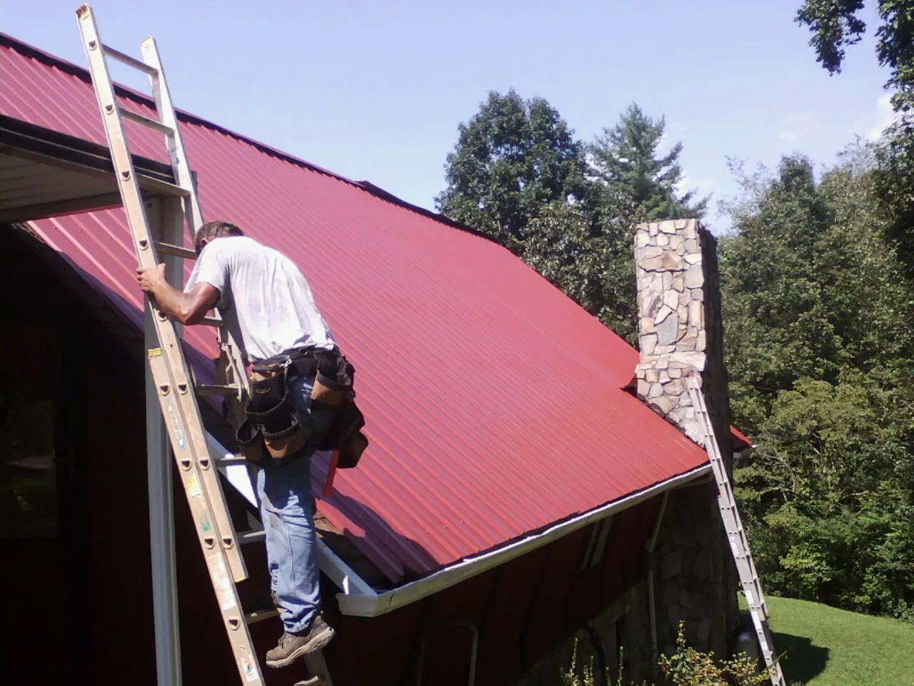 Roofer on a ladder beside a red metal roof and stone chimney — Soffit & Fascia Repair by Roofing Brothers in North Reading